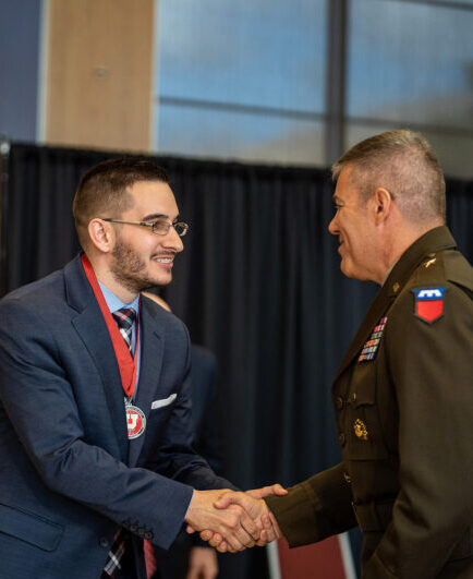 DSC9335_2000-682×532-1-e1700081335468 a decorated military official shakes the hand of someone wearing a medal with the U logo on it