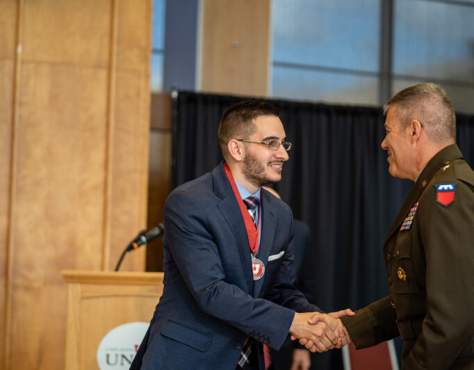 DSC9335_2000-682×532 someone shaking hands with someone else. One is wearing a medal with the U logo, the other is wearing military medals